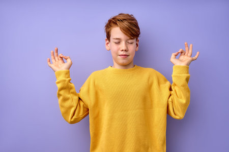 Portrait of young teenage boy keep calm, meditating, isolated on purple studio background. Kid in...