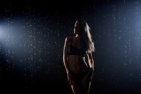 beautiful young lady posing under drops of water, isolated on black studio background. portrait of sexy woman in black swimsuit underwear is illuminated by light, having athlete bodyの写真素材