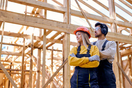 team of constructor engineers in New wooden residential construction home framing. Construction site. Beautiful caucasian lady and man in workwear, hardhat and headset looking at side, at sunny dayの写真素材