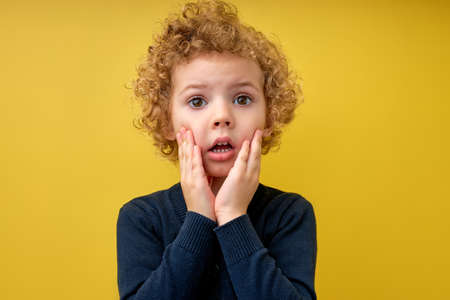 Portrait of surprised kid boy in shock, touching cheeks, stand with widely opened mouth and eyes, caucasian child looking at side, saw something, isolated on yellow studio background. copy spaceの写真素材