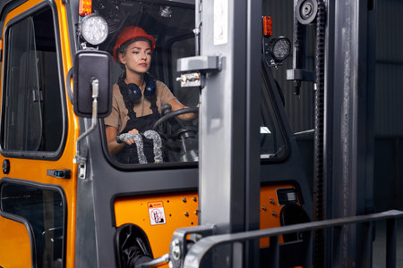focused Female worker driving loader in industrial container warehouse, looking confident, dressed...