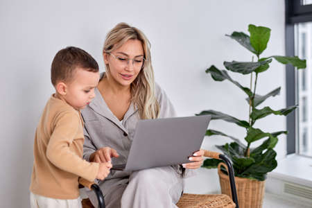 lovely caucasian family woman and little son using laptop computer together in bright cozy apartment at home. caucasian blonde lady work on laptop while child boy stand next to her.の写真素材