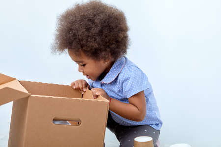 multiracial small girl with funny expression is looking for toy in carboard box isolated on white studio background. child in blue shirt with fluffy curly hair, want to play, looking inside of boxの写真素材
