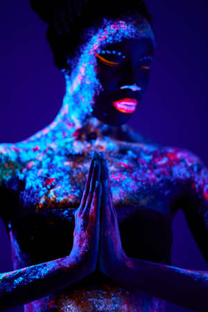 Fashion model woman in neon light, model with fluorescent make-up in prayer position, Art design of female posing in UV, colorful make up. Isolated on dark blue background. pray for the bestの写真素材