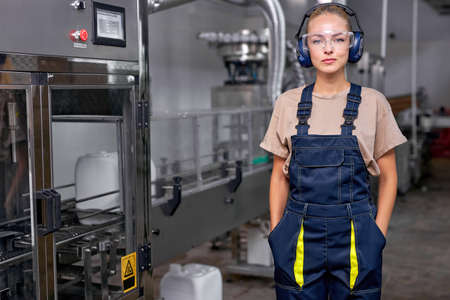 young industrial woman engineer in spectacles standing in factory at work place, dressed in blue uniform. pesticides production in factory. lady engineer look at camera, posing confidentlyの写真素材