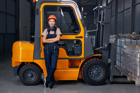 smiling woman labor worker near forklift driver in industry factory logistic shipping warehouse, posing with arms folded, dressed in uniform and orange protective hardhat, after successful good jobの写真素材