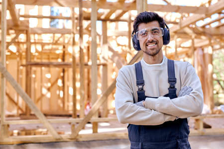 smiling caucasian man in headset and uniform at work place, wooden house. Bearded young male in protective spectacles look at camera holding arms folded. architecture, renovation, constructionの写真素材