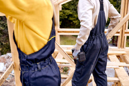 cropped male holding drill tool in hands, working with female contractor on building site, working together, help each other, unrecognizable male female in work clothesの写真素材