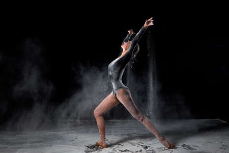 gorgeous slender ballet dancer woman wearing bodysuit, posing dancing among the cloud of flying flour on black studio background, make performance. Artistic, commercial, monochrome design.の写真素材