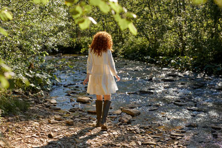 Rear View On Curly Redhead Lady Having Rest Relaxing On River, Walking Alone At Sunny Day Or Evening, View From Back On Caucasian Female In White Romantic Dress. In Forest, Nature Countrysideの写真素材