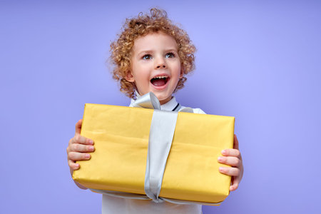 child boy holding yellow gift box in hands, celebrating birthday, get presents by relatives and parents, friends. cheerful kid of caucasian appearance with curly hair looking up with opened mouthの写真素材