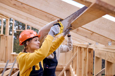 caucasian confident female contractor in orange helmet using saw hand cutting wooden beam in wooden house site. Young responsive man builder help and support to saw board.の写真素材