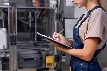 cropped female worker checking robotic line for bottling and packaging pesticides into bottles and canisters, take notes in folder-tablet , writing with pen on paper. production, manufacturingの写真素材
