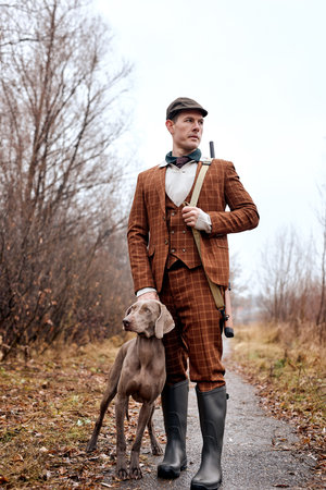 Caucasian Hunter man with rifle shotgun and with hunting dog Weimaraner walking in rural road during...