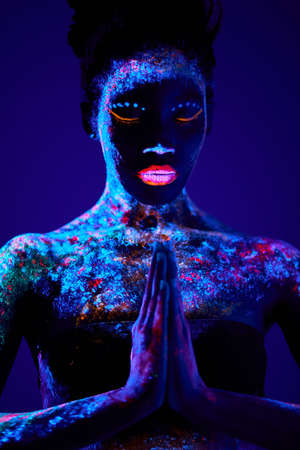 beautiful sensual woman of black appearance in fluorescent paint makeup, posing in prayer pose. luminescence paint, body art, neon lights. isolated on dark background. portrait, indoorsの写真素材