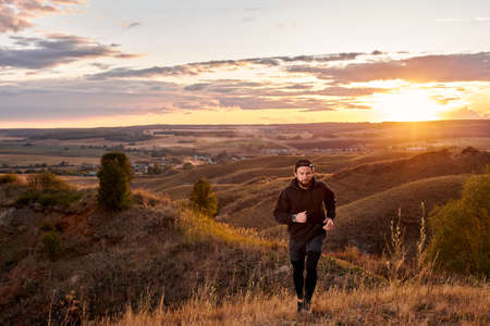 caucasian man jogging along the field, wearing sportive clothes, practice running outdoors in the morning at sunrise, in nature. young male engaged in sport, motivated. strong and healthy personの写真素材