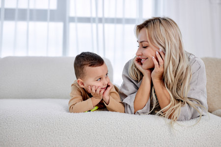 woman cuddling talking to small kid son, having fun together on sofa in studio living room. Smiling...