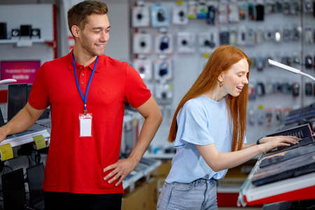 caucasian woman is looking for the best laptop in computer department of electronics store. Lady buyer chooses laptop in modern technology store, nice male consultant help customer with choiceの写真素材