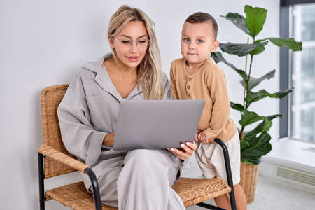 beautiful caucasian family woman and little son using laptop computer together in bright cozy apartment at home. caucasian blonde lady work on laptop while child boy stand next to her.の写真素材