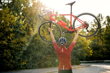 View back on Sporty man in sportive clothes, protective helmet and glasses carrying raising bike up after successful competition, posing at camera with red bike outdoors. sport and active lifestyleの写真素材