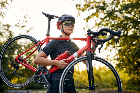 athlete caucasian competitor cyclist carrying bicycle in hands, in nature on road, wearing sportive outfit, in glasses and helmet. sport, fitness, workout, training, healthy lifestyle conceptの写真素材