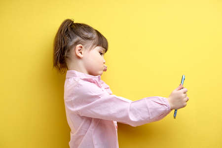 portrait of child girl taking photo or making video on smartphone, posing at phones camera, kissing. Adorable little girl in pink blouse using modern technologies. Children and human lifestyleの写真素材
