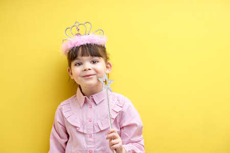 happy smiling girl with crown on head, holding magic wand, looking at camera, yellow studio wall, copy space. little fairy posing, having caucasian appearanceの写真素材