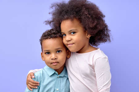 portrait of two curly adorable siblings brother and sister hugging each other, isolated on purple studio background. portrait of black birl and boy in casual wear posing looking at cameraの写真素材