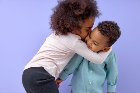 Two cheerful black american siblings, sister kissing her brother, isolated on purple background....