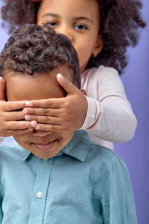 cheerful carefree little girl covering eye of boy with arms, sincerely smiling, laughing, having fun and playing peek a boo game. indoor studio shot isolated on purple background.focus on boyの写真素材