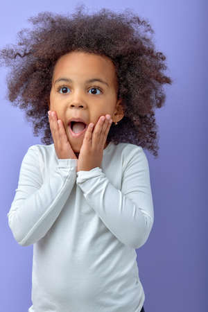 Photo of black little kid amazed shocked omg wow fake novelty reaction isolated over purple color background. emotional curly black child girl in casual shirt look at side with surprised expressionの写真素材