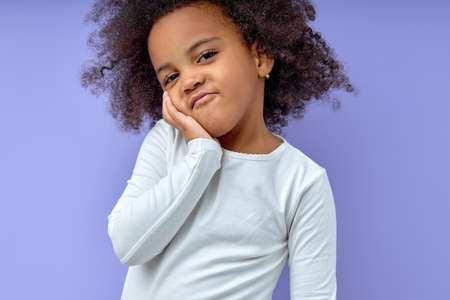 young funny curious black kid girl holding hands on cheeks looking contemptuously standing on isolated purple background, listening to someone, thinking. childrens emotions conceptの写真素材