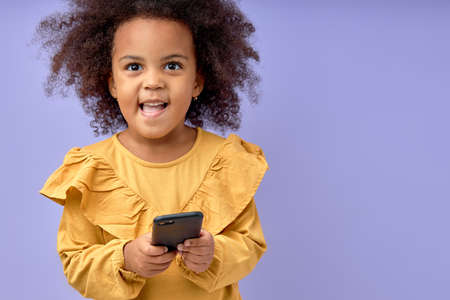Pretty Child Girl With Curly Hair Using Smartphone, Look At Camera Smiling, Isolated On Purple Studio Background, Kid in Yellow Dress or Blouse Posing, Interested On Game, Cartoon. Portraitの写真素材
