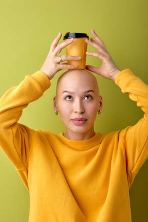 Confident hairless female have fun holding cup of coffee on bald head, enjoy life. European woman with allopecia disease laughing, dressed in casual yellow shirt. Isolated green studio backgroundの写真素材