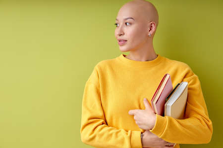bald young european american woman looking at side holding books in hands, alopecia and cancer concept, copy space. satisfied hairless female in casual yellow outfit, education and universityの写真素材