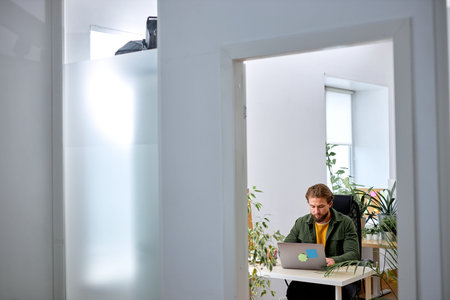 Analyzing data on laptop. young concentrated bearded man in casual wear sit with laptop behind desk...