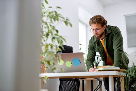 Bearded young confident guy stand behind office desk with laptop thinking working alone. handsome...
