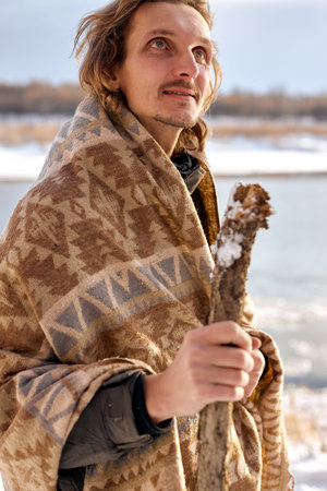 active tourist with wooden stick looking around while walking in winter forest on cold day, wrapped...