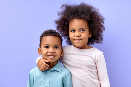 portrait of two adorable siblings brother and sister hugging each other, isolated on purple studio background. portrait of black birl and boy in casual wear posing looking at cameraの写真素材