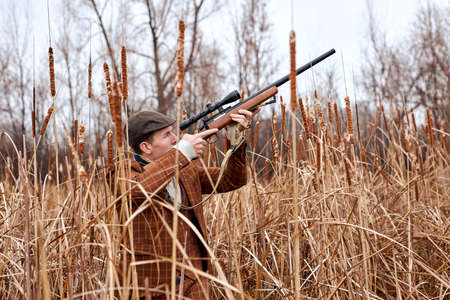 european caucasian hunter is hunting in forest, looking through the rifle, soft focus. in countryside. male in brown suit confidently aiming rifle to the side, concentrated on hunting.の写真素材