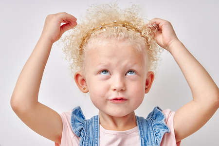 Kid girl touching amazing blonde curly hair, looking thoughtful. Isolated on white background, copy space. portrait of caucasian child in denim dress looking up, funny face. children emotionsの写真素材