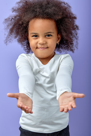 black cute girl stretches arms for a hug, kind adorable child smiling, isolated on purple studio background. portrait of cheerful curly kid in white shirt posing at camera, charming kidの写真素材