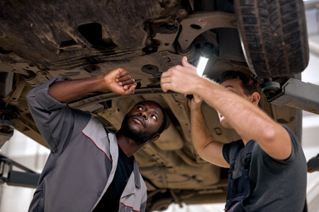 two diverse Handsome mechanics in uniform examining car while working in auto service. young men in overalls concentrated on work together, using lamp, under car. vehicle, transport, car serviceの写真素材