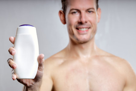 close-up portrait of excited guy showing bottle of gel or shampoo, for advertisement, isolated on gray studio background. young male of caucasian appearance smiling, focus on bottleの写真素材