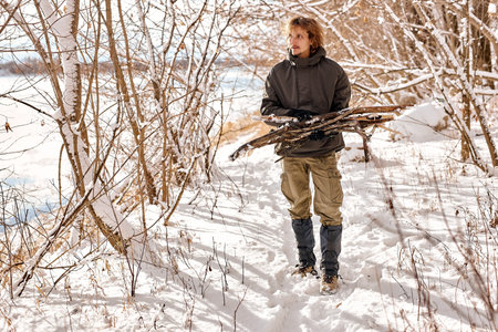young caucasian male gathering wood for fire in forest, nature. lonely guy in warm clothes in snowy...