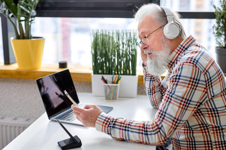 Handsome elderlycaucasian man sitting alone, using modern laptop and headset, bearded male having...