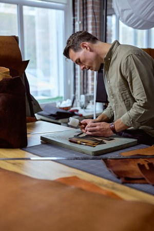 young handsome man focuses on making brown handmade leather wallet. Sewing wallet. Piercer punches...