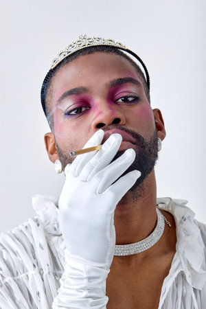 young transgender with bright night party makeup smoking cigarette posing isolated over neutral white background. Black american guy in white shirt, with earrings, in crown. Lgbtq, beauty conceptの写真素材