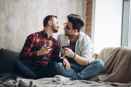 European male gay couple drinking wine sitting on the soft plaid in the room with loft interior.の写真素材