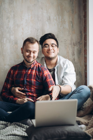 European homosexual male couple enjoying leisure time together, sitting by table and watching online movie on laptop computer in the living room with monochrome interiorの写真素材